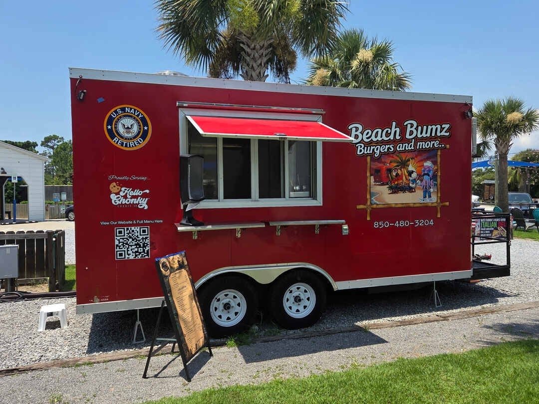 Red food truck with Beach Bumz logo and serving window, parked on grass with palm trees behind it
